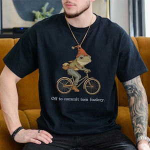 Könnte beinhalten: Schwarzes T-Shirt mit einer Illustration eines Frosches auf einem Fahrrad, der einen Hut trägt und eine Tasche dabei hat. Der Text "Off to commit tom foolery" ist darunter gedruckt. Das T-Shirt ist aus weichem Material.