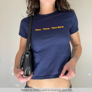 May include: Navy blue crop top with the text "They / Them / That Bitch" in yellow. The t-shirt has short sleeves and a crew neck. A black purse is hanging from the shoulder.