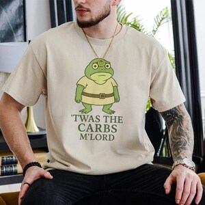 May include: Beige t-shirt with a cartoon frog wearing a yellow shirt and brown belt. The text "'TWAS THE CARBS M'LORD" is printed below the frog. The shirt is a casual style.
