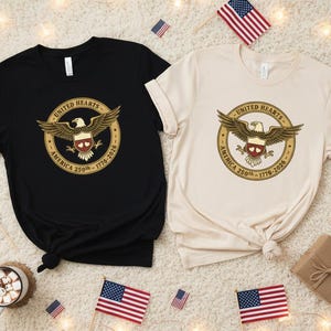 May include: Two t-shirts, one black and one cream, each with a circular graphic featuring a golden eagle, the words "United Hearts America 250th 1776-2026", and a red and white shield. American flags and a mug of hot chocolate are in the background.