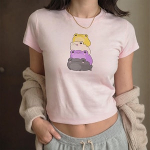 Op de afbeelding: Een lichtroze crop top met een ontwerp van cartoonkikkers in geel, paars en grijs. De kikkers zijn op elkaar gestapeld. De top heeft korte mouwen en een ronde hals.