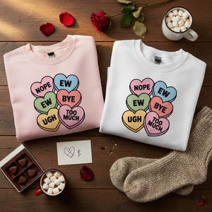 Può includere: Due felpe, una rosa e una bianca, ciascuna con una grafica di caramelle a forma di cuore. Le caramelle dicono "NOPE", "EW", "BYE", "UGH" e "TOO MUCH". Su una superficie di legno.