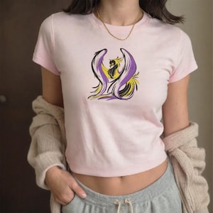 May include: A light pink crop top featuring a stylized phoenix design in black, yellow, and purple. The short-sleeved shirt is made of soft material and is a casual wear item.