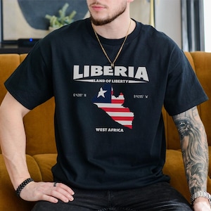 May include: Black t-shirt with the word "LIBERIA" in white block letters above a map of Liberia in red, white, and blue. The text "LAND OF LIBERTY" is above the map, and "WEST AFRICA" is below it. Coordinates are also included.