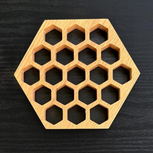 以下が含まれることがあります： 六角形の木製コースター。ハニカム模様のデザインで、明るい茶色の木材を使用。六角形の切り抜きが連なっており、蜂の巣を連想させるデザインです。