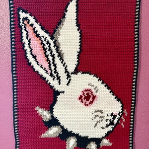 May include: Crocheted wall hanging depicting a white rabbit head with a black and grey studded collar against a red backdrop. The rabbit has pink inner ears and a red eye. The border is black with grey stitching.