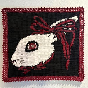 以下が含まれることがあります： 黒い背景に赤いリボンが付いた白いウサギの頭が描かれたかぎ針編みの正方形。ウサギは赤と灰色の目を持っています。ボーダーはスカラップの赤いトリムです。この作品はおそらく手作りです。