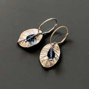 May include: Silver leaf earrings with a dark blue teardrop-shaped bead hanging from each leaf. The earrings have a simple hoop design.