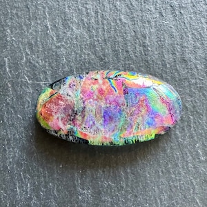 May include: A colorful iridescent glass oval with a textured surface. The glass has shades of pink, purple, blue, and green.
