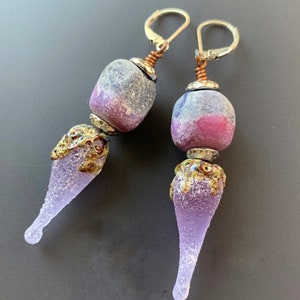 May include: A pair of dangle earrings with a purple and pink glass bead and a textured purple glass teardrop pendant.