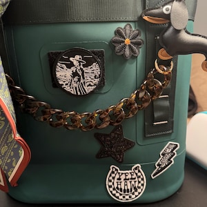 May include: Dark green cooler bag adorned with patches, a chain, and a dachshund charm. Patches include a cowgirl, a star, a daisy, a lightning bolt, and a horseshoe with "Yee Haw" text. The chain is tortoise-shell colored.