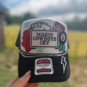 May include: A black and white trucker hat with the text "MAKIN COWBOYS CRY". The hat features a red rose, playing cards, an 8-ball, and a lightning bolt. The hat has a foam front and a snap closure.