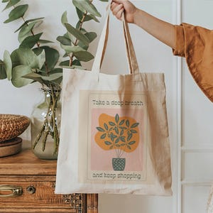 Puede incluir: Una bolsa de tela beige con un diseño gráfico que presenta una planta en maceta y el texto "Respira hondo y sigue comprando".