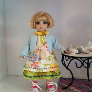 May include: A doll with blonde hair and blue eyes wearing a light blue cardigan over a floral dress and pink trainers. The doll is sitting at a small black table with a teapot and teacups.