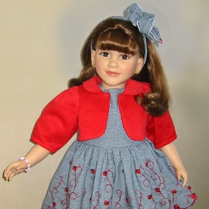 Könnte beinhalten: Eine Puppe trägt eine rote Jacke über einem blau-weiß karierten Kleid mit roten Stickereien. Die Puppe hat braunes Haar und trägt ein weißes Stirnband mit einer blauen Schleife.