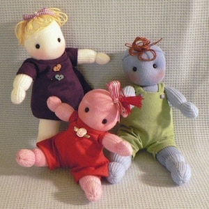 Puede incluir: Tres muñecas de calcetín hechas a mano. Una muñeca viste un vestido morado con botones en forma de corazón y un lazo amarillo. Otra muñeca lleva un atuendo rojo con una rosa. La tercera muñeca lleva un mono verde y tiene un peinado de hilo marrón.