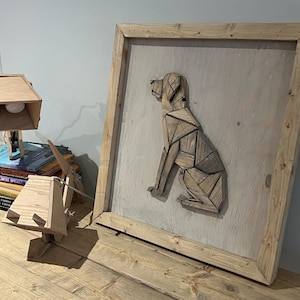 May include: A wooden table with a stack of books, a wooden lamp, and a framed wooden dog sculpture. The dog is made of various pieces of wood and is sitting in a profile pose. The frame is made of natural wood and has a light gray background.