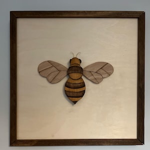 May include: A wooden bee cutout in a brown frame. The bee is made of layered wood and has a brown and yellow striped body with brown wings.