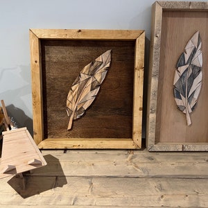 Puede incluir: Dos piezas de arte de pared de madera con diseños de plumas geométricas. La pieza de la izquierda tiene un fondo marrón y una pluma marrón claro. La pieza de la derecha tiene un fondo marrón claro y una pluma gris y blanca.