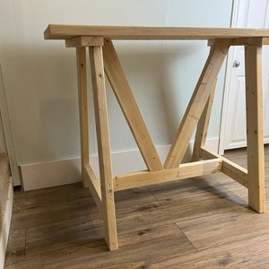 May include: A wooden table with a simple, rustic design. The table has a rectangular top and a base made of four legs that are connected by a series of cross beams. The table is made of unfinished wood and has a natural wood color.