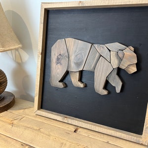 Puede incluir: Una escultura de oso de madera en un marco marrón sobre un fondo negro. El oso está hecho de múltiples piezas de madera, cada una con un patrón de veta diferente. El marco está hecho de madera natural con un acabado rústico.