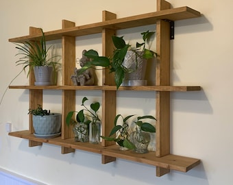 May include: A wooden wall shelf with multiple shelves and compartments, decorated with potted plants and a small figurine. The shelf is made of natural wood with a light brown finish.