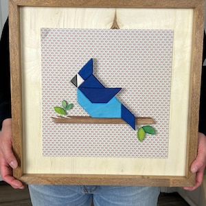 May include: A framed artwork featuring a geometric blue jay perched on a branch with green leaves. The bird is composed of various shades of blue. The background has a patterned design. The frame is made of wood.