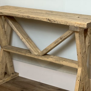 May include: A rustic wooden console table with a natural wood finish and a unique X-shaped base. The table has a wide, rectangular top and a simple, farmhouse style.