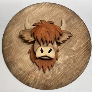 May include: A wooden wall hanging featuring a Highland cow's head. The cow's face is made of layered wood, with a light brown snout, dark brown fur, and cream-colored horns. The cow is mounted on a round, stained wooden plaque.