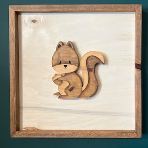 May include: A wooden framed wall decor featuring a brown squirrel holding a nut. The squirrel is made of wood and is attached to a light brown wood background.