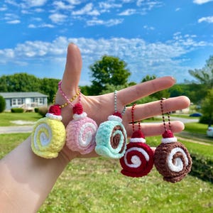 Puede incluir: Se muestran cinco adornos de rollo de pastel de ganchillo en amarillo, rosa, turquesa, rojo y marrón. Cada uno tiene un remolino blanco y un pequeño sombrero rojo. Cuelgan de cadenas de cuentas. El fondo es un cielo azul brillante.