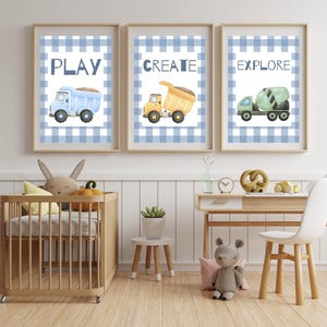 May include: Three framed wall art prints featuring watercolor construction vehicles on a blue and white gingham background. The prints display the words PLAY, CREATE, and EXPLORE above a blue dump truck, a yellow dump truck, and a green cement mixer.