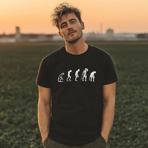 Peut inclure: T-shirt noir à col rond avec un motif blanc représentant l'évolution d'une créature. Le motif montre une série de figures, commençant par une forme voûtée et progressant vers un extraterrestre humanoïde. Le t-shirt est en matière souple.
