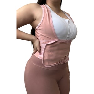 May include: A pink waist trainer with adjustable straps and a zipper closure. The waist trainer is worn over a white sports bra.