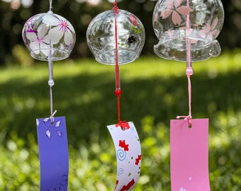 May include: Three clear glass wind chimes with floral designs and colorful paper tags. The chimes hang from red and white strings against a blurred green background. The chimes have different floral patterns in pink and white.