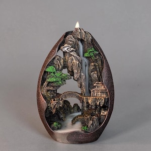 Op de afbeelding: Decoratieve tafel fontein met een miniatuur Aziatisch landschap. De fontein heeft een waterval, brug en klein gebouw, alles in een bruin, eivormig frame. Het water stroomt over de rotsen, wat een serene sfeer creëert.