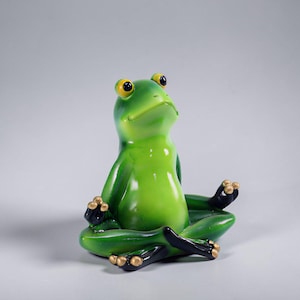 May include: A green frog ornament in a seated yoga position. The frog has yellow eyes, black feet with gold detailing, and is set against a light grey backdrop. The figurine is a decorative item.