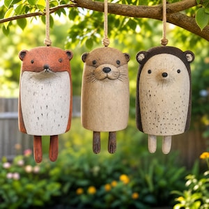 May include: Three hanging animal-shaped wind chimes featuring a fox, an otter, and a hedgehog. Each figure has a cylindrical body, small ears, and dangling legs, suspended by a thick jute rope. The fox is orange and white, the otter is tan, and the hedgehog is dark brown and cream with speckled details.