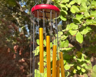 May include: A hanging wind chime with a red wooden top and gold-colored metal tubes. Black strings connect the top to the tubes. The background is blurred green foliage.