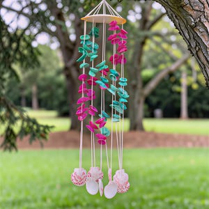 May include: A decorative wind chime featuring teal and magenta shell-shaped beads suspended from a circular top. The chime hangs from white strings, with seashell pendants at the base. The background shows a blurred green lawn and trees.