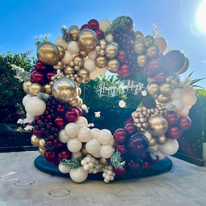 May include: A large, circular balloon arrangement in gold, burgundy, white, and silver. The balloons are arranged in a wreath shape, with the words "Happy Holidays" in the center. Greenery is interspersed throughout.