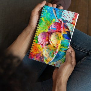 May include: A colourful spiral notebook featuring a watercolour painting of ice cream in a bowl. The ice cream is various shades of pink, yellow, and blue with colourful sprinkles. The background is a vibrant yellow, blue, and green.