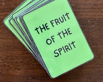 Puede incluir: Una pila de tarjetas rectangulares con esquinas redondeadas. Cada tarjeta tiene un fondo verde claro con texto negro: "THE FRUIT OF THE SPIRIT". Las tarjetas están apiladas sobre una superficie de madera marrón oscuro.
