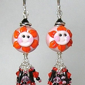 May include: A pair of handmade dangle earrings. Each earring features a pink bead with an orange flower design and a smiling face. Below, a cluster of red, black, and purple beads hangs from silver wire, creating a whimsical and colorful accessory.