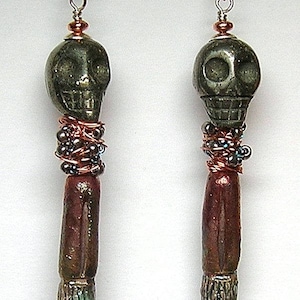 May include: A pair of handcrafted earrings featuring a skull-shaped bead in a dark, metallic green hue. The skull is connected to a long, cylindrical bead with a dark red-brown color, accented with copper wire and small beads. The earrings have a unique, artistic design.