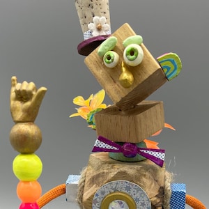 Puede incluir: Una escultura de robot caprichosa y artesanal. El robot presenta una cabeza de madera con ojos saltones, un sombrero de copa de corcho y una mano dorada que sostiene un bastón con cuentas de colores. Tiene brazos naranjas y zapatos negros.