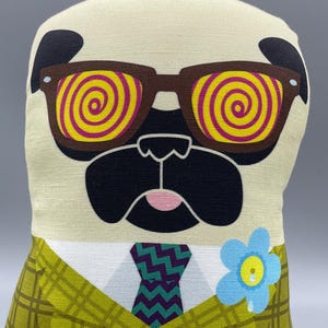 Puede incluir: Muñeco de peluche de un pug con gafas de sol marrones con lentes en espiral amarillas y rosas. El pug viste una blazer a cuadros verdes, una corbata estampada y un broche de flor azul. El muñeco tiene una nariz negra y una lengua rosa.