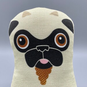 May include: A beige plush toy shaped like a pug with brown eyes, a black nose, and a brown beard. The toy is sitting on a brown and white patterned fabric.