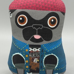Puede incluir: Un pug de peluche gris que lleva un sombrero rojo y una chaqueta vaquera azul con una camiseta negra que dice "IMOTOR IMPERFECT CYCLES".
