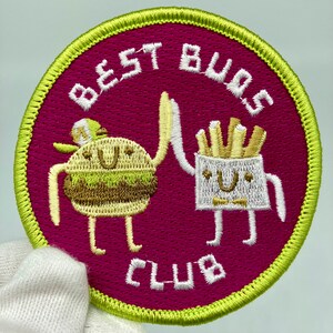 May include: Embroidered patch with a magenta background and a green border. The patch features cartoon illustrations of a hamburger and a french fries container giving a high-five. The text "BEST BUDS CLUB" is embroidered in white.
